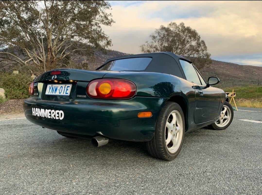 Dark green Mazda convertible parked on a road with "HAMMERED" sticker on the rear bumper.
