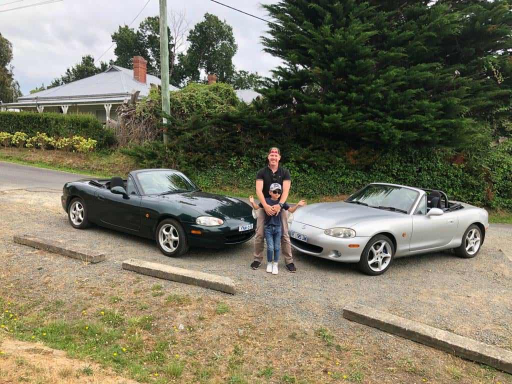 Two convertible cars parked on a gravel area with a man and child standing between them.