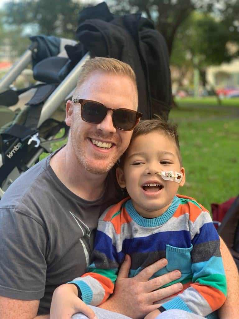 Smiling man wearing sunglasses hugs happy child with a cheek bandage outdoors near stroller.