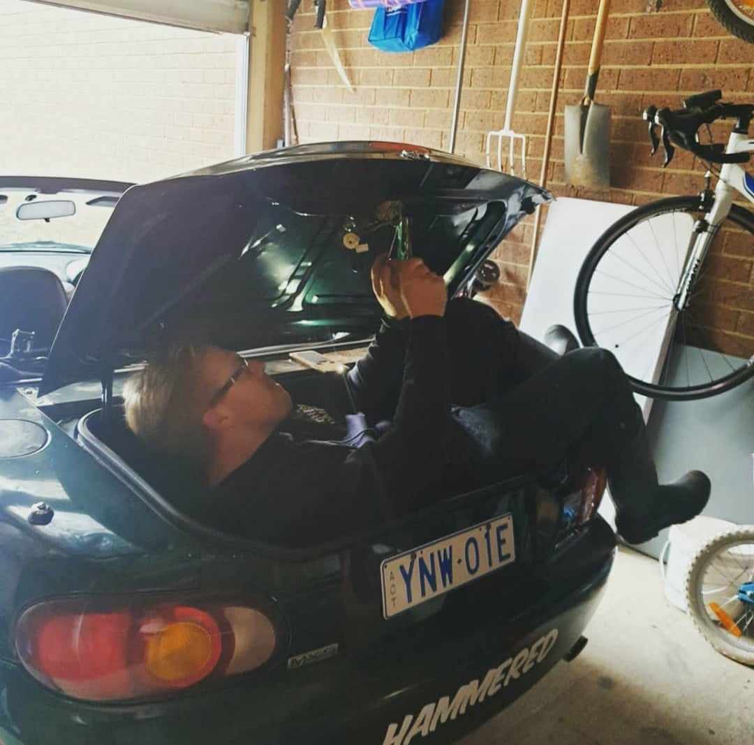 Person lying inside a car trunk with the lid open, holding a bottle, in a garage.