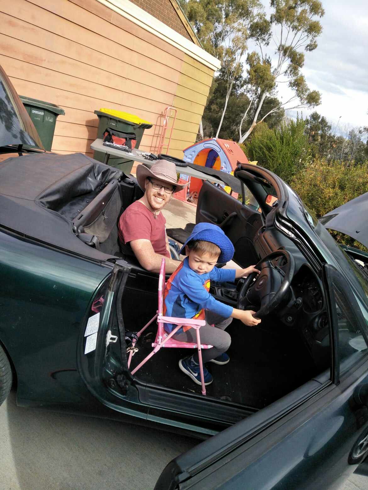Child in blue hat sitting on pink chair in driver's seat of convertible car, adult beside him.