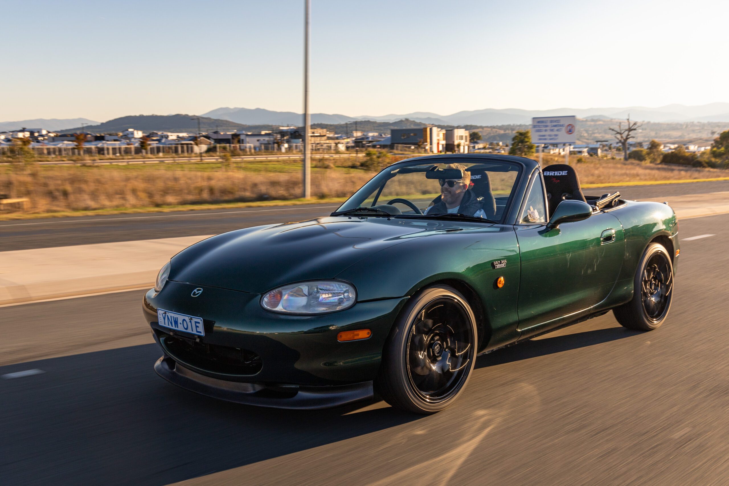 Dark green Mazda convertible driven by a man on a sunny road with mountains in the background.