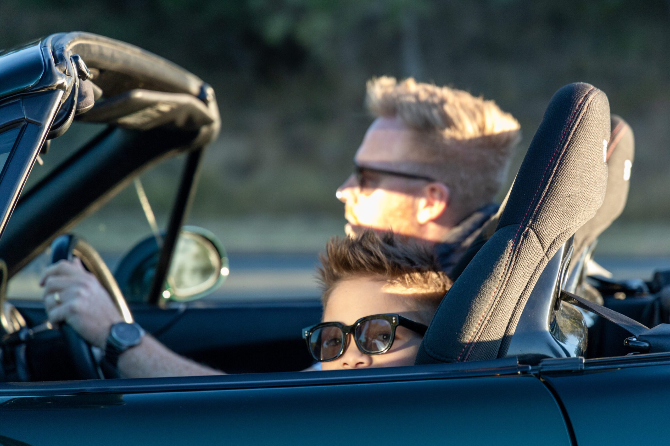 Child wearing sunglasses peeks over car door as adult drives a convertible with the top down.