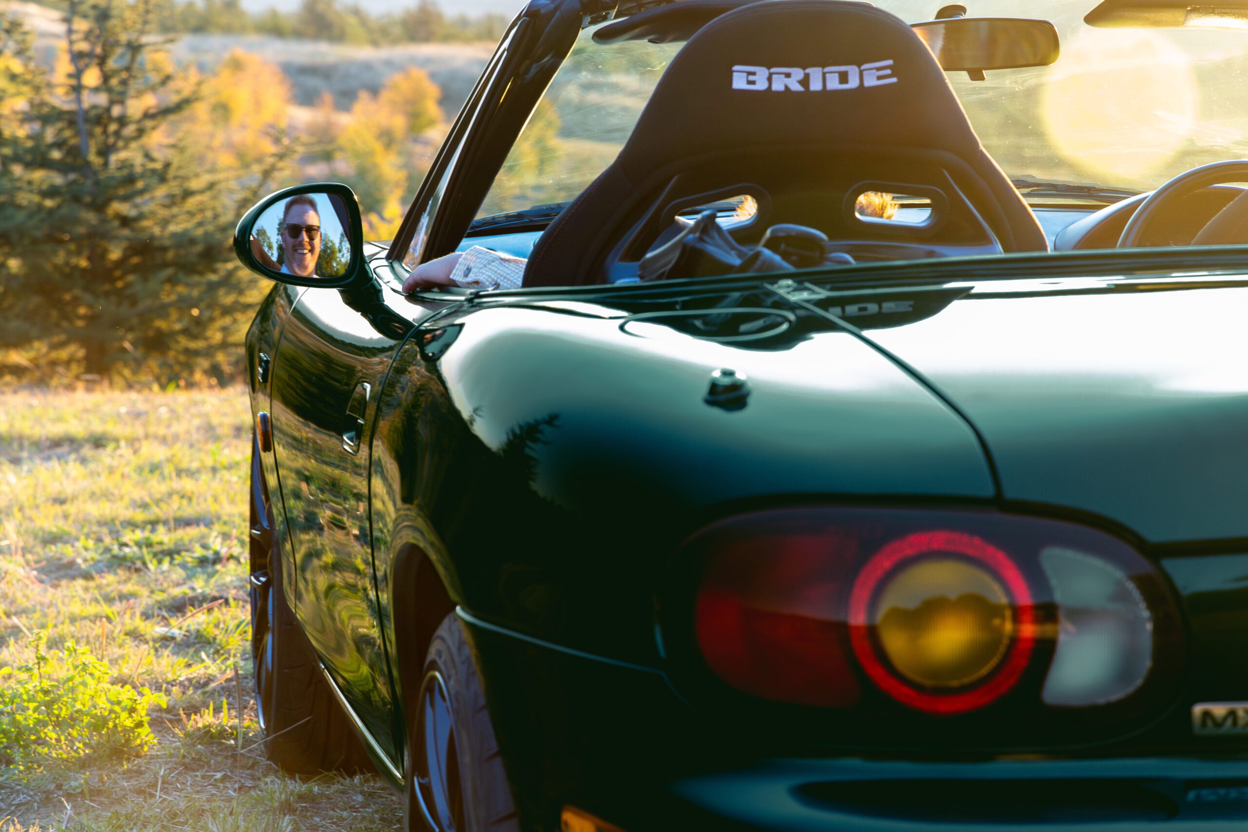 Man with sunglasses reflected in side mirror of green convertible car during sunset.