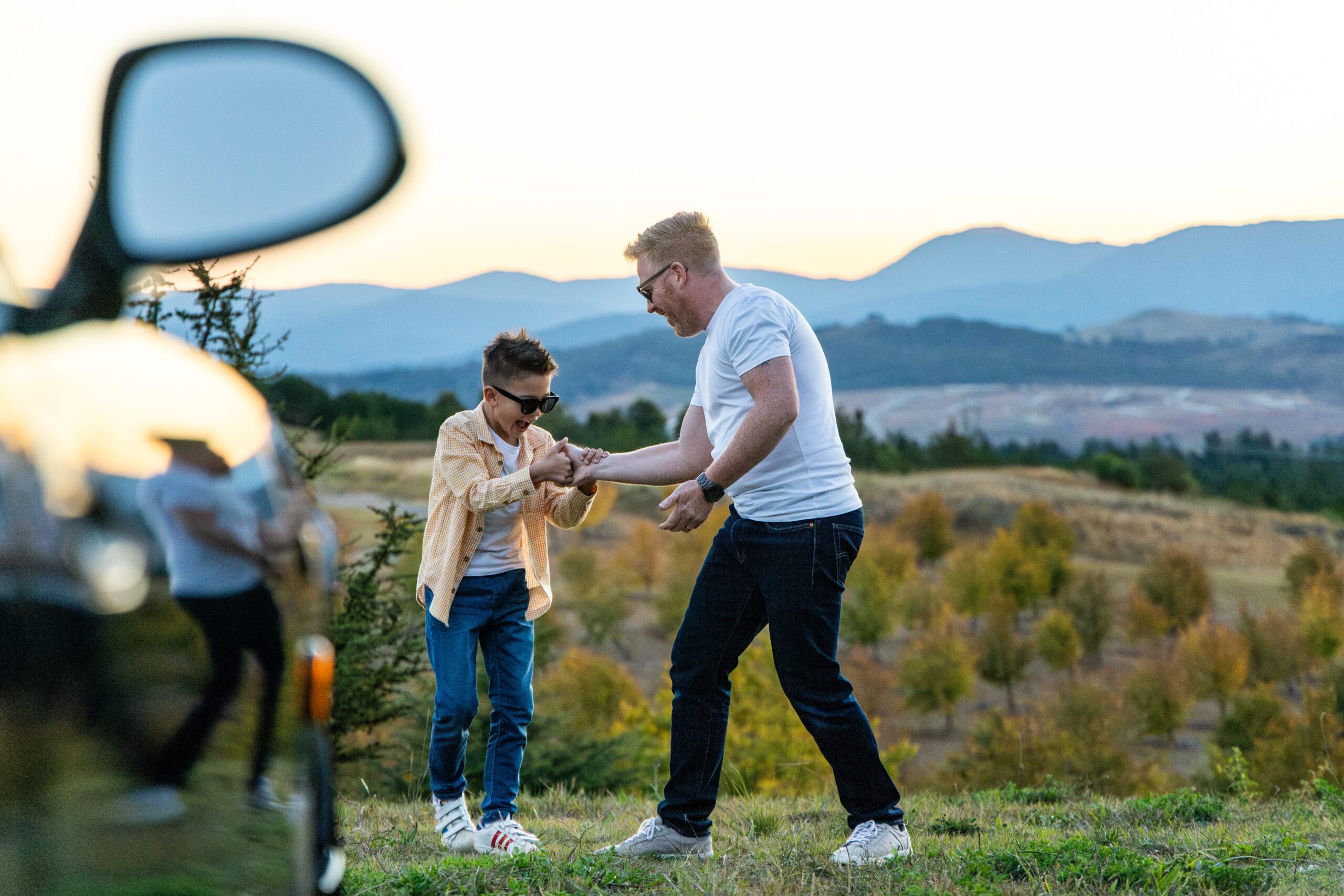 Father and son wearing sunglasses laughing and playing outdoors near a car at sunset.