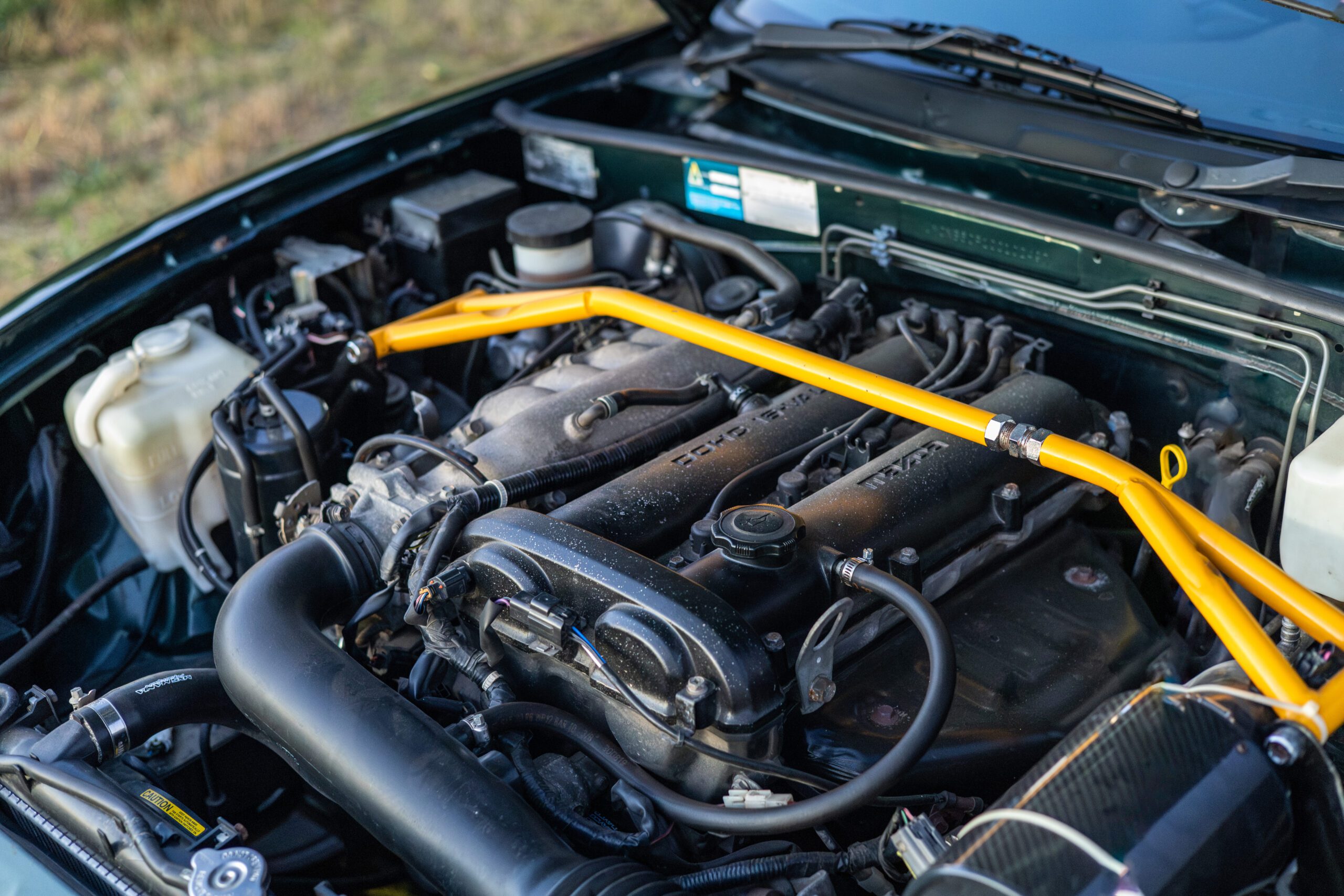 Car engine bay with a yellow strut bar and various hoses and components visible.