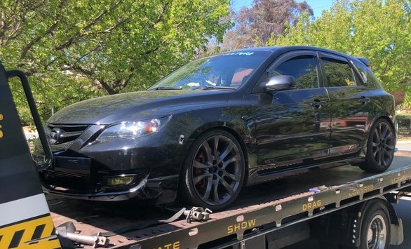 Black sporty hatchback car loaded on a flatbed tow truck with trees in the background.