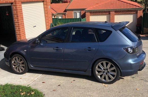 Blue hatchback car parked on a driveway next to a brick house with a closed garage door.