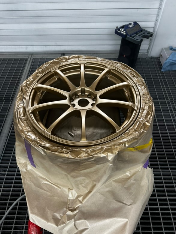 Gold-colored car wheel rim partially wrapped in protective paper on a grated floor.