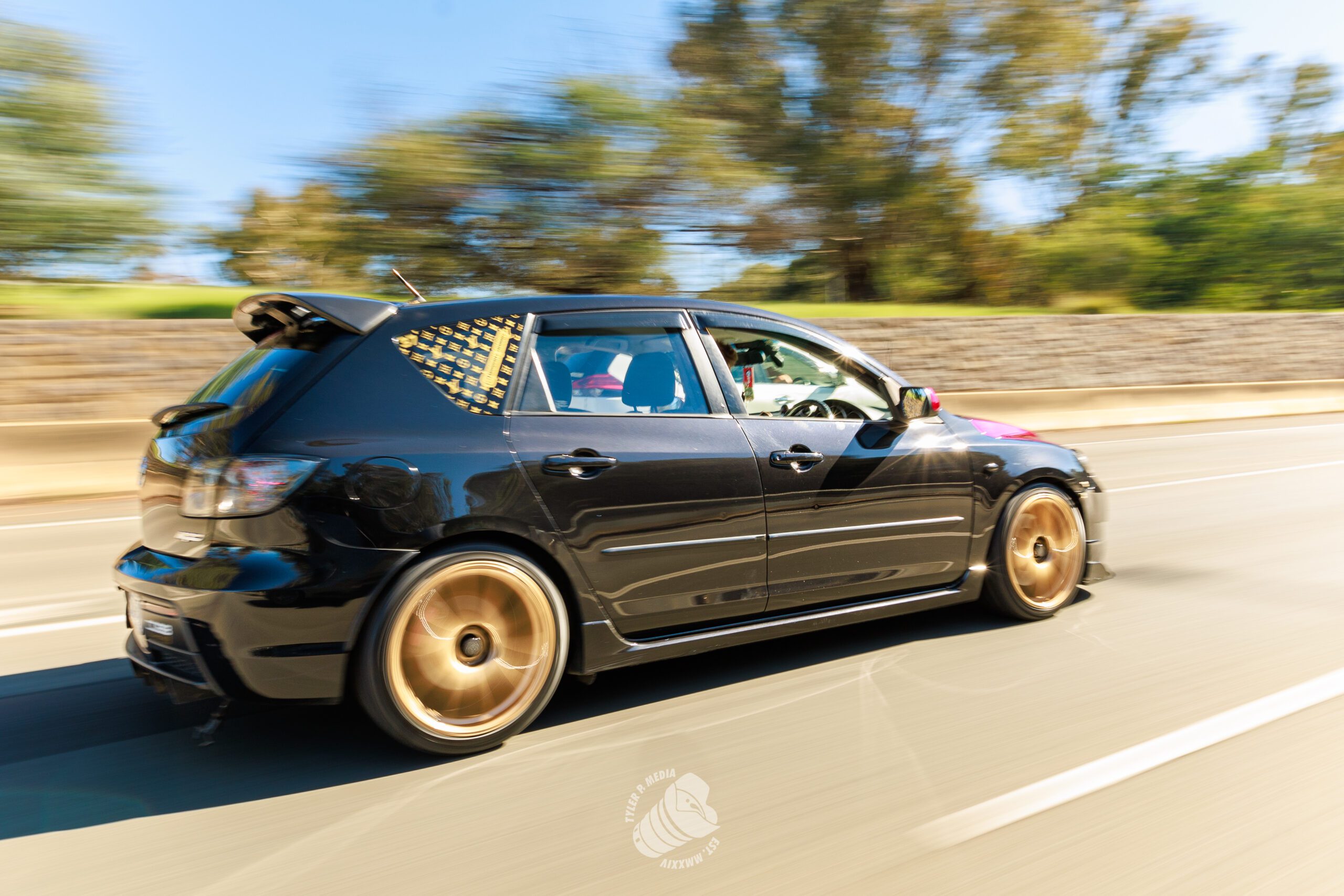 Black hatchback car with gold rims and patterned rear window driving fast on highway.
