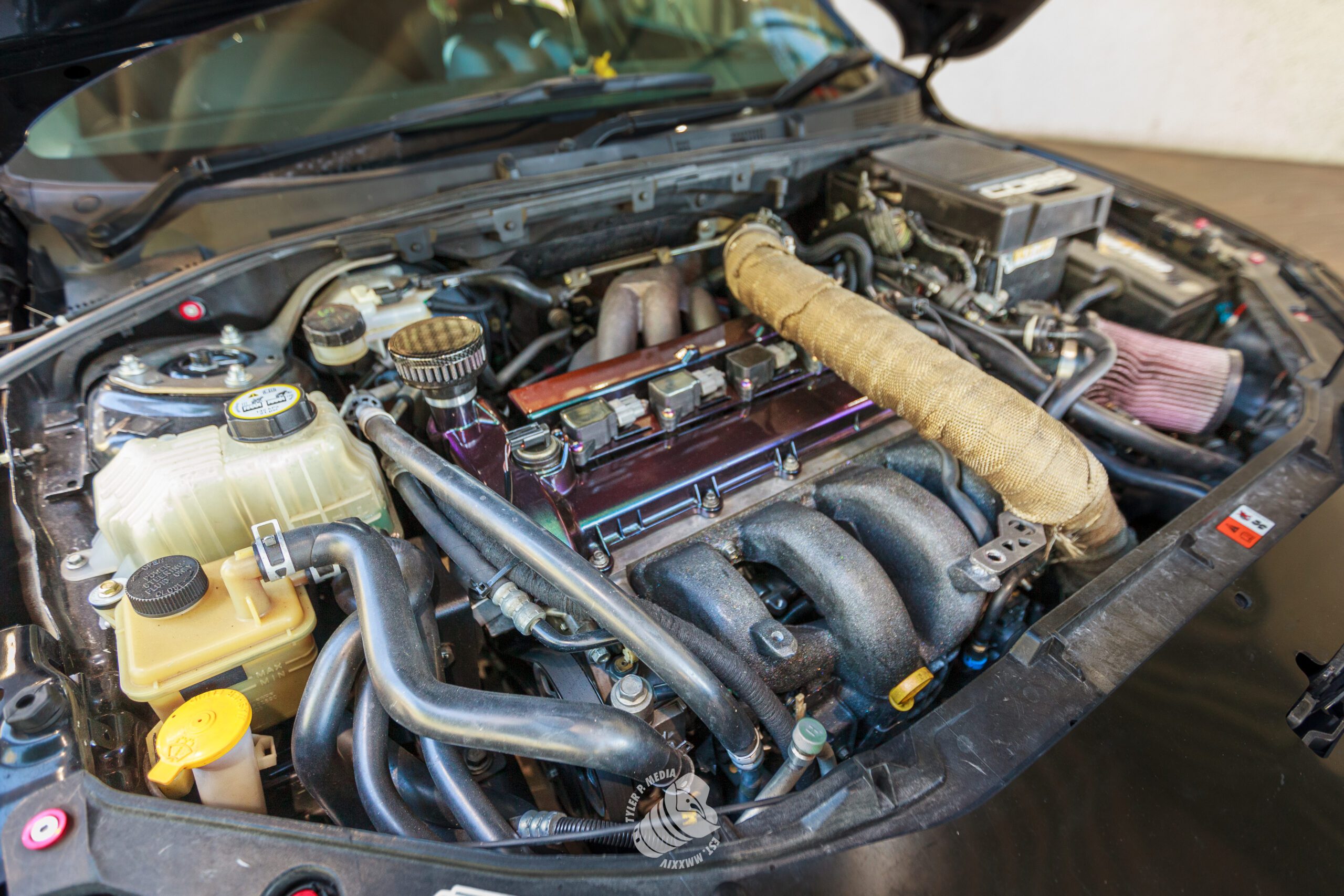 Car engine bay with various components, hoses, and a wrapped exhaust manifold visible.