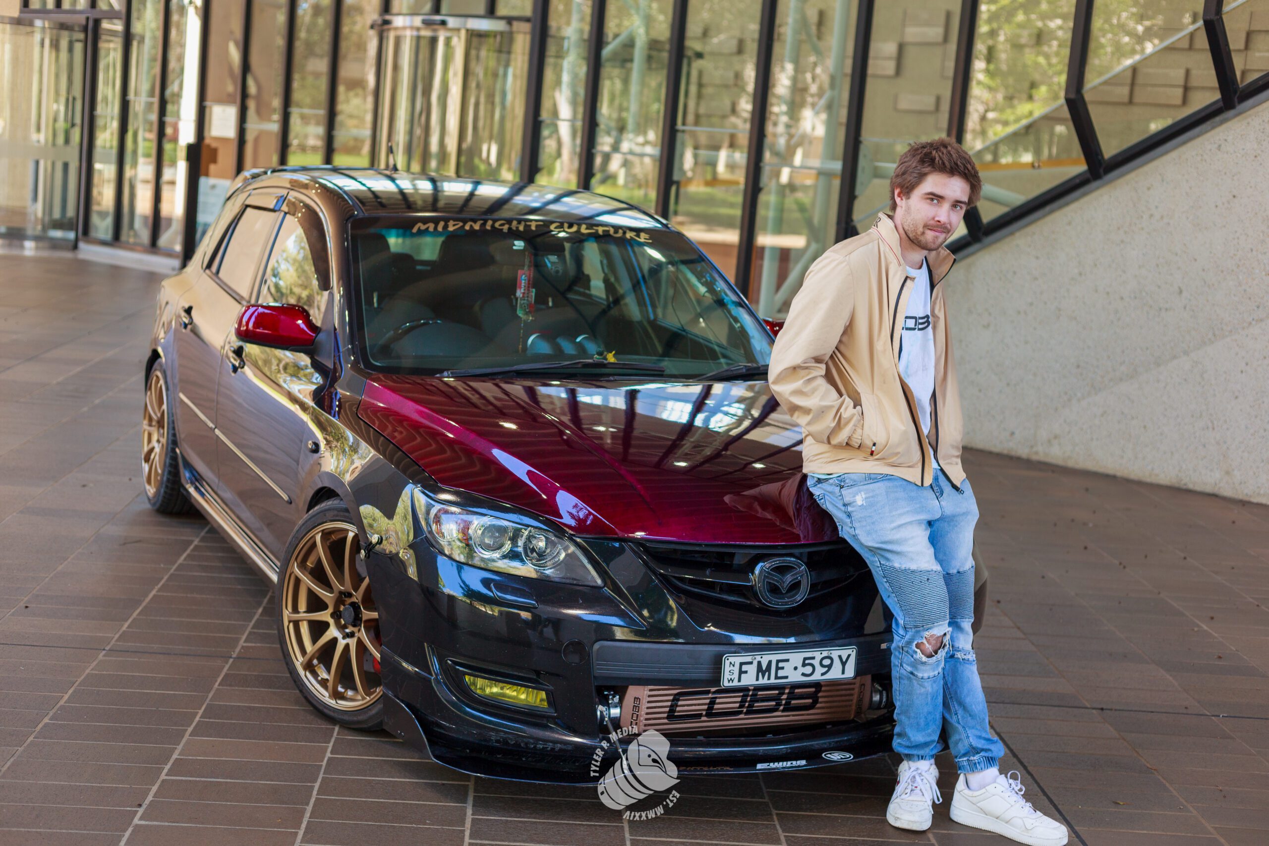 Young man in casual wear leaning against a shiny black and red Mazda car with gold rims.