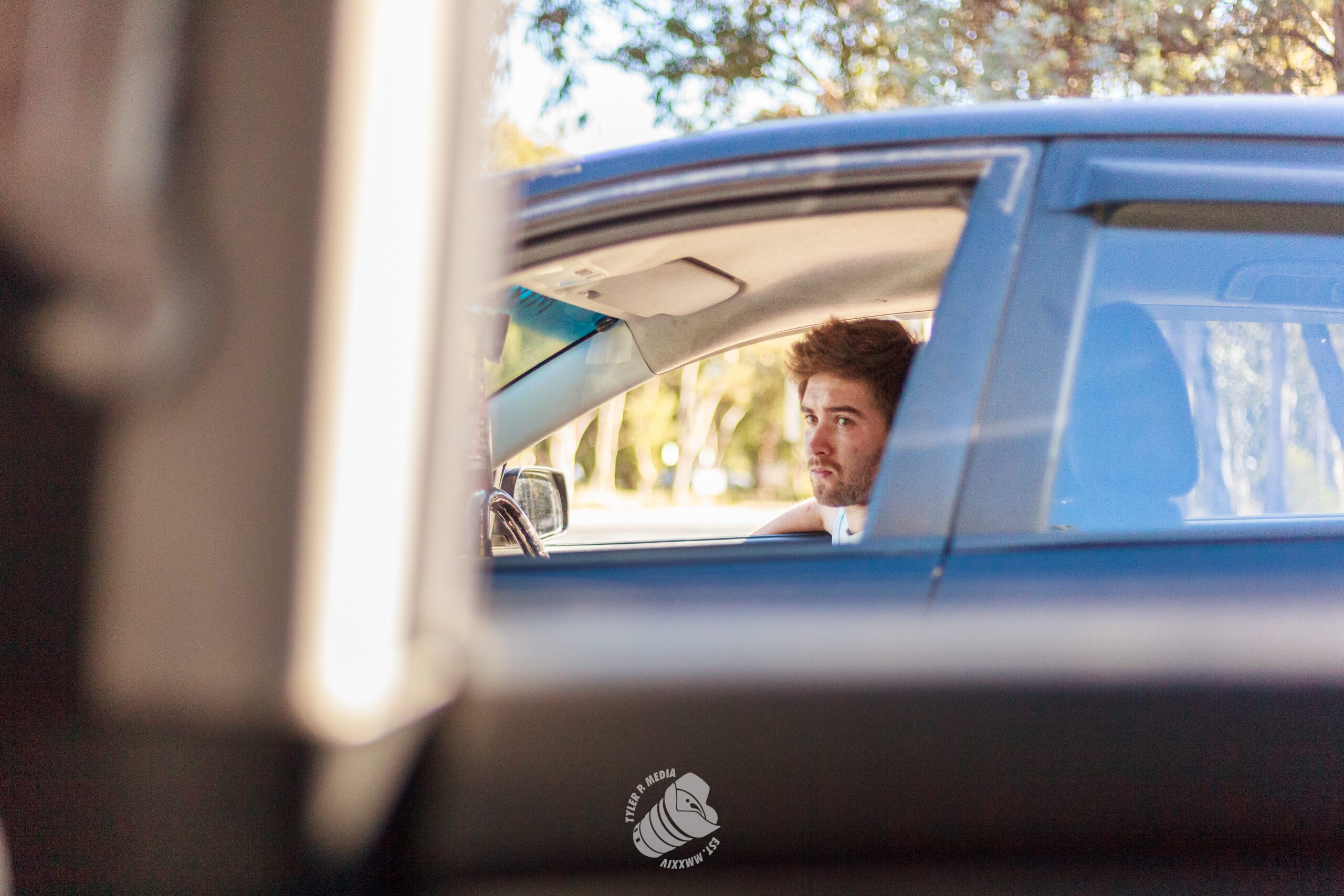 Man with beard sitting inside a blue car, looking out the driver's side window during the day.
