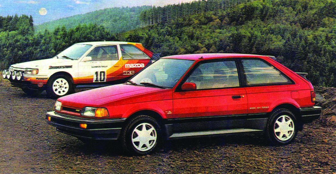 Two vintage hatchback cars, one red and one white with racing decals, parked on a gravel road with trees in background.