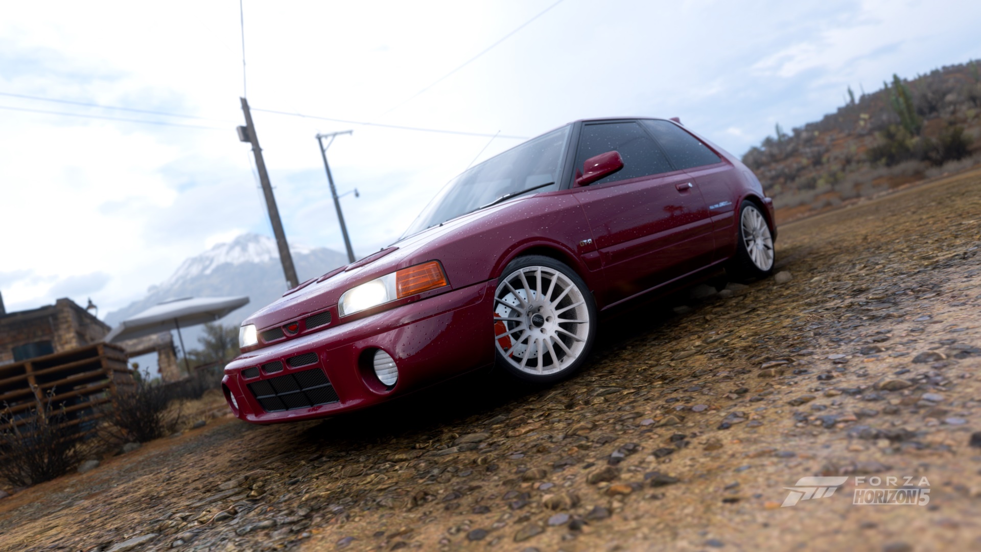 Maroon hatchback car with white wheels on a gravel road, with mountains and buildings in the background.