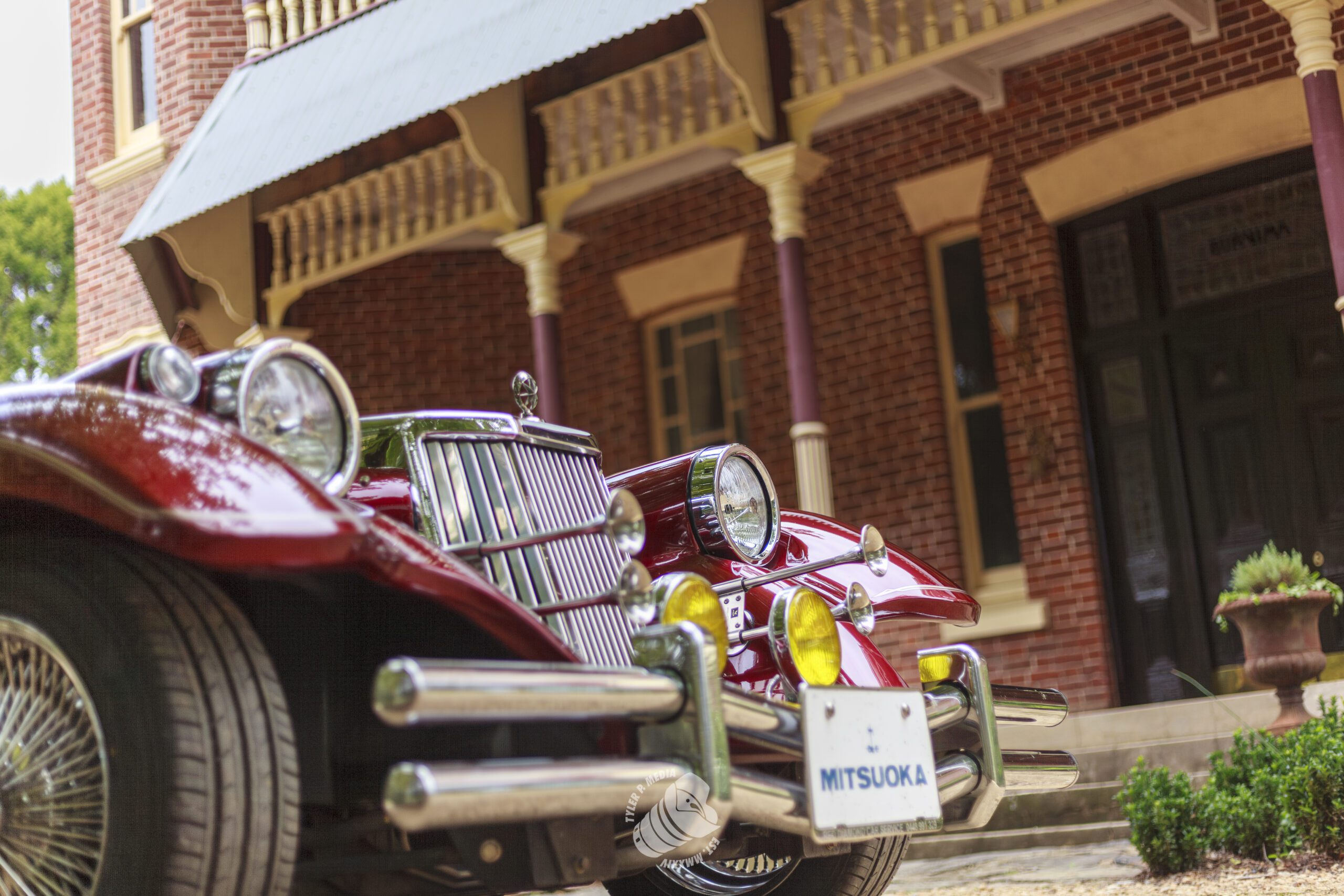 Close-up of a red Mitsuoka vintage car parked in front of a brick building with pillars.