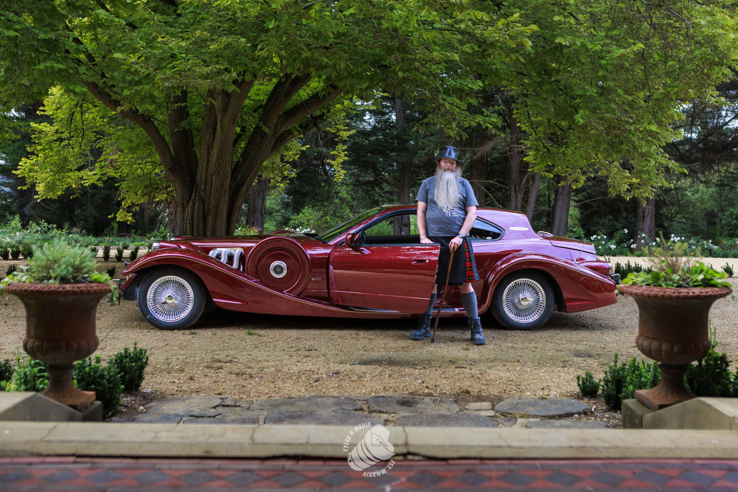 A man with a long beard, wearing a hat and kilt, stands with a cane beside a vintage red car.
