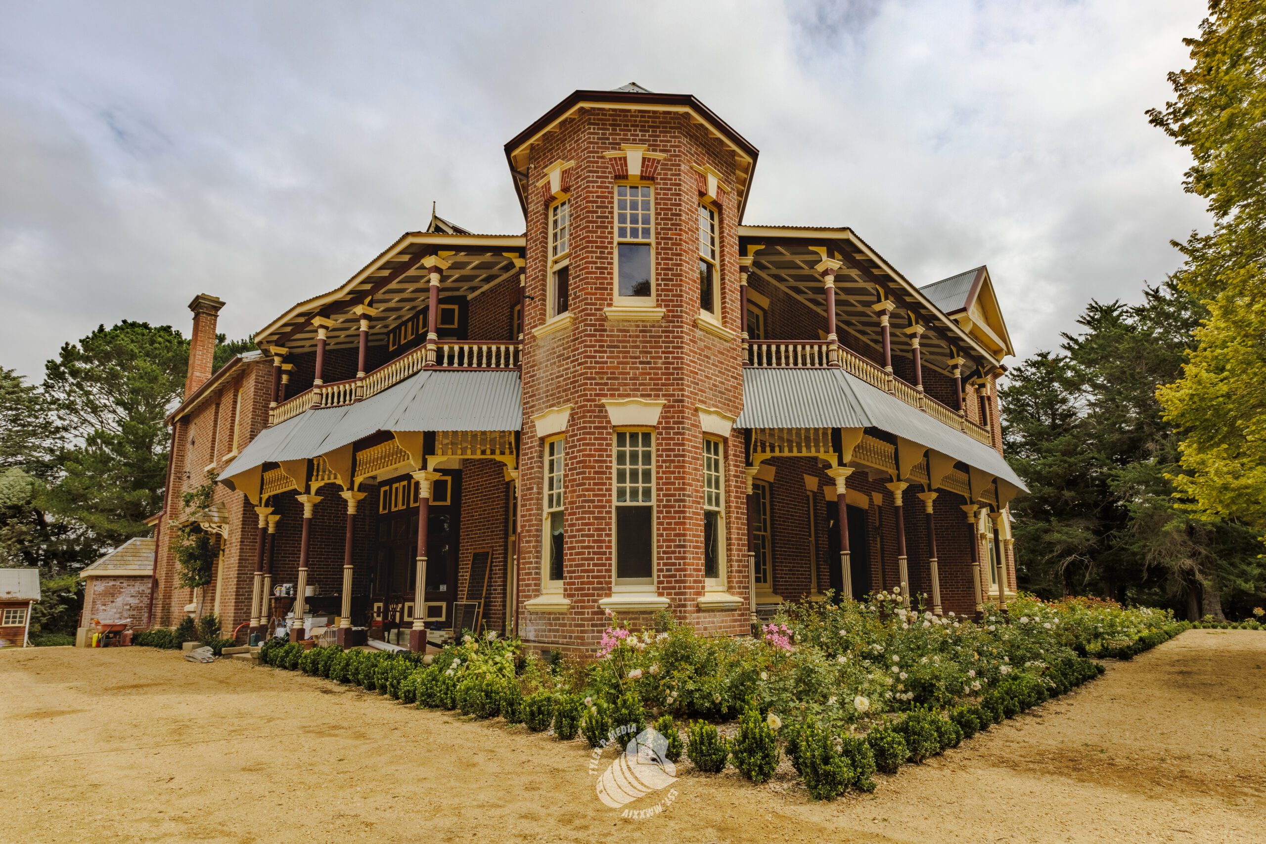 Large two-story brick house with wraparound porch and manicured garden under cloudy sky.