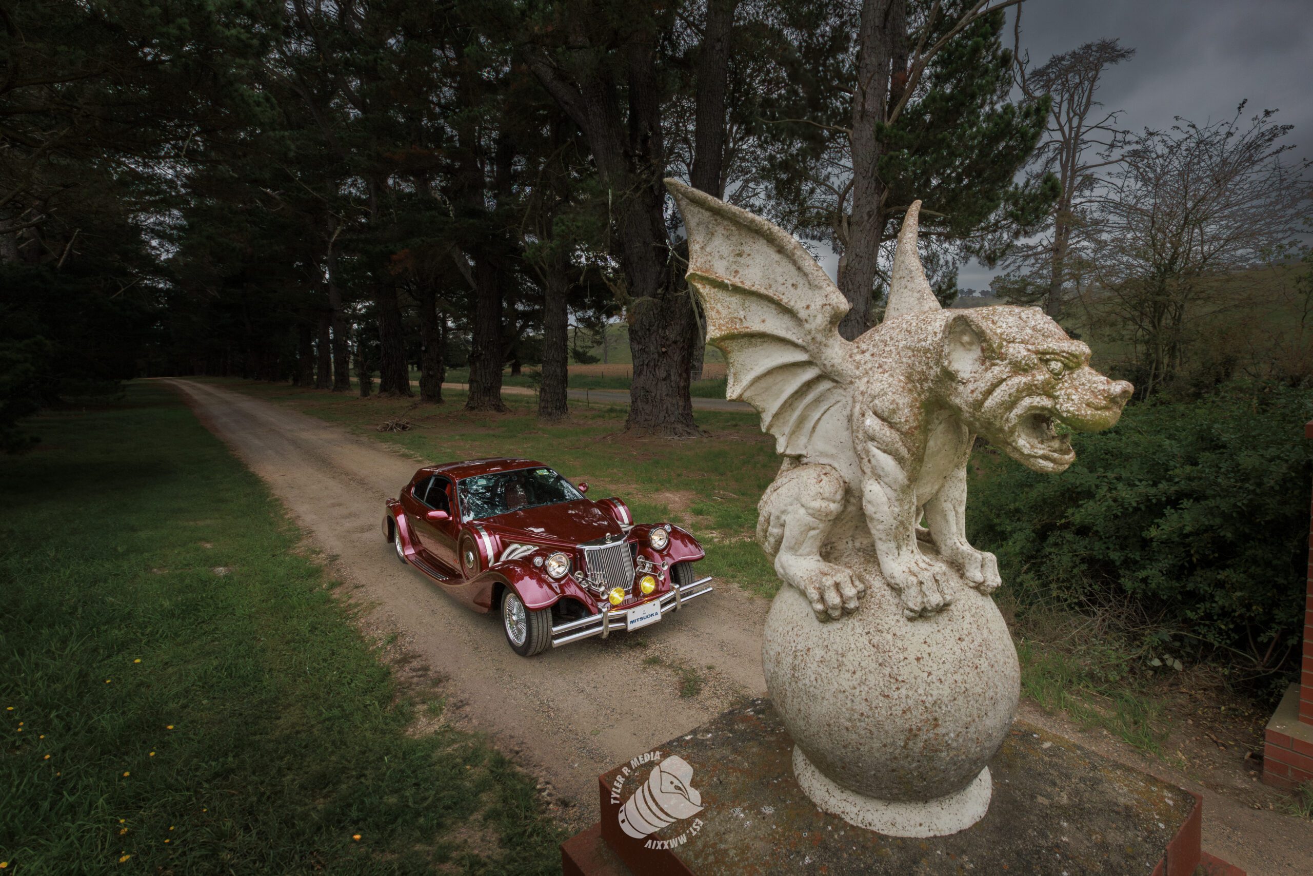 Vintage red car driving on a forest road past a stone gargoyle statue on a cloudy day.