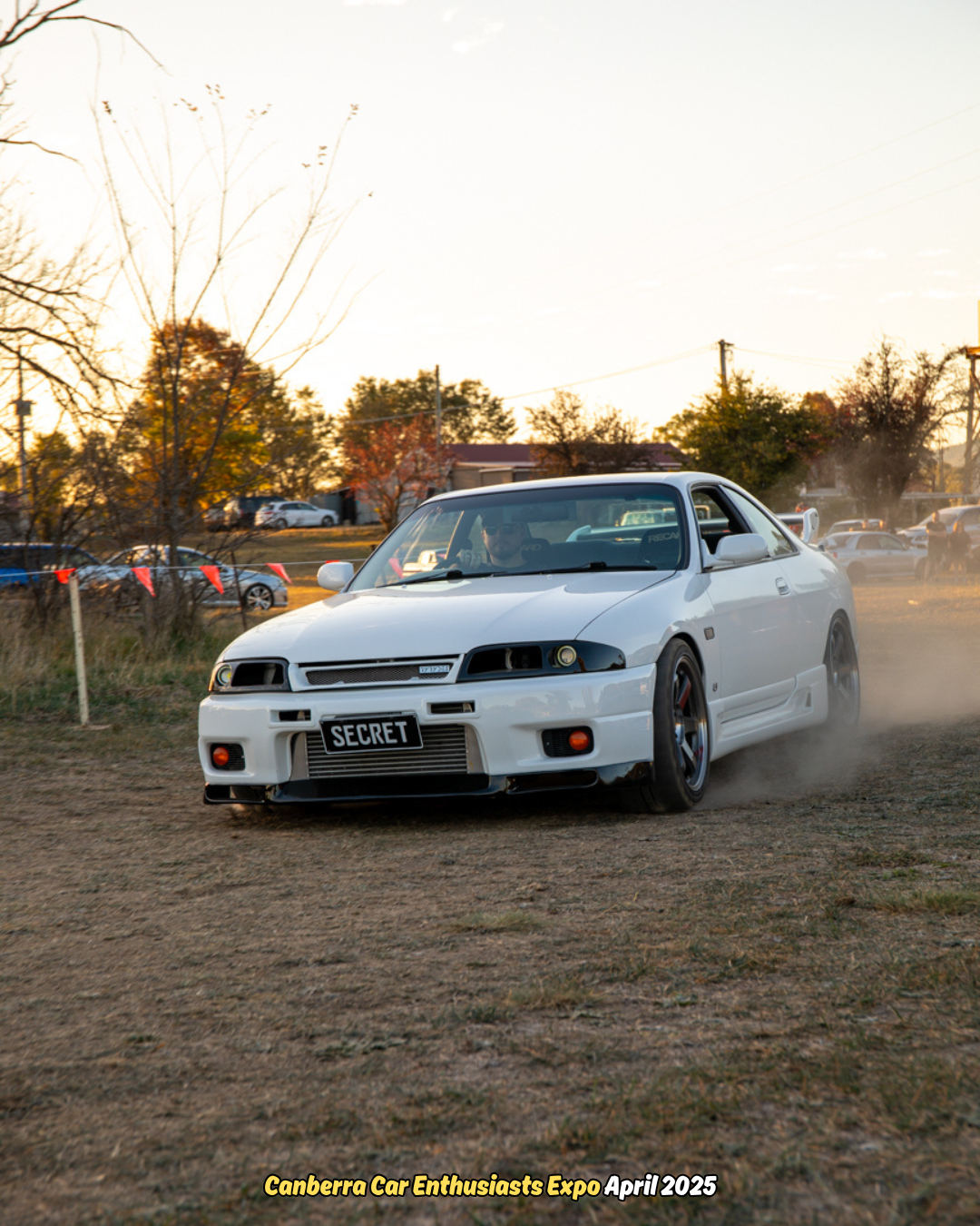 White sports car drifting on dirt at an outdoor car expo with dust swirling behind it.