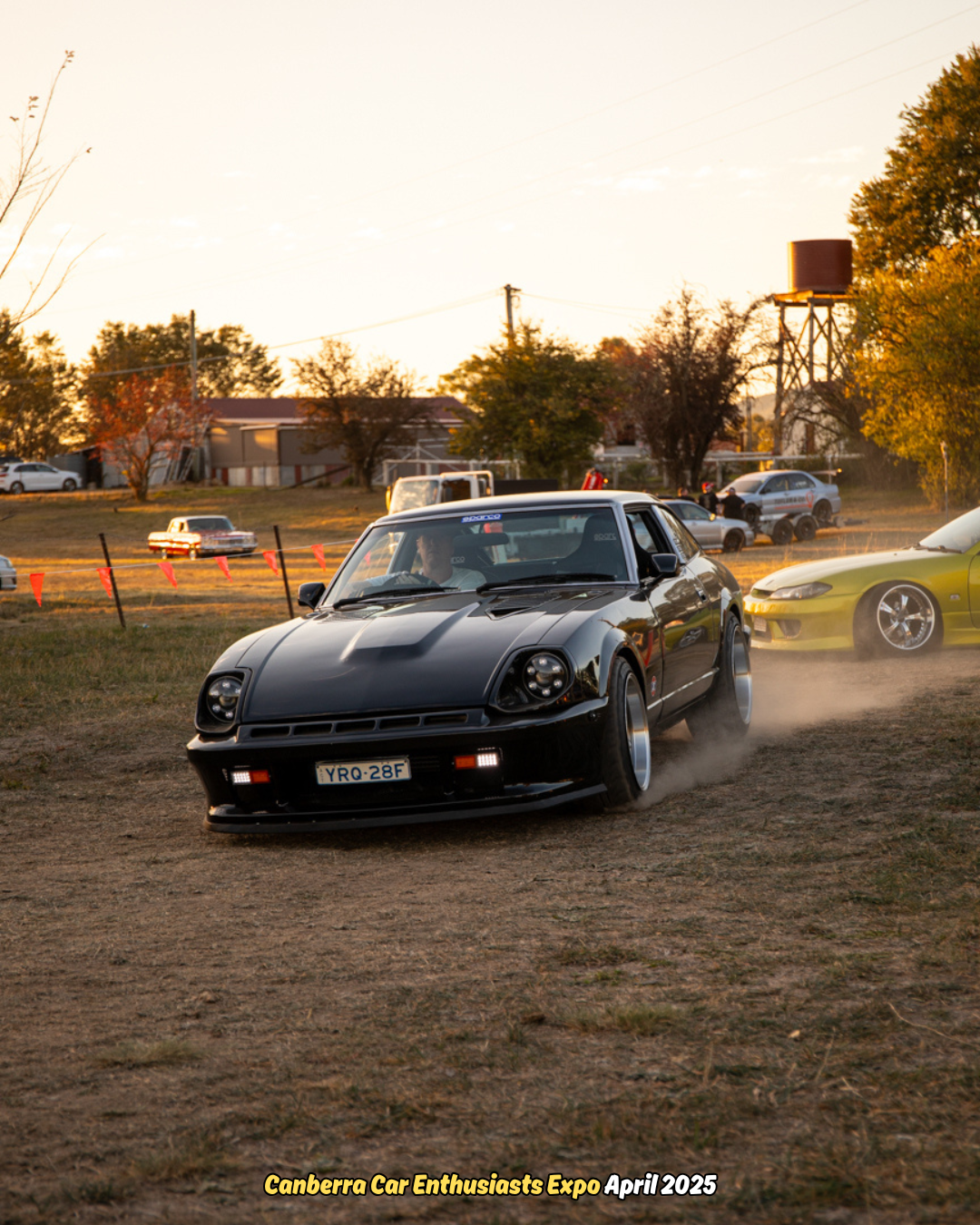 Black sports car drifting on dirt with dust rising, trees and other cars in background.