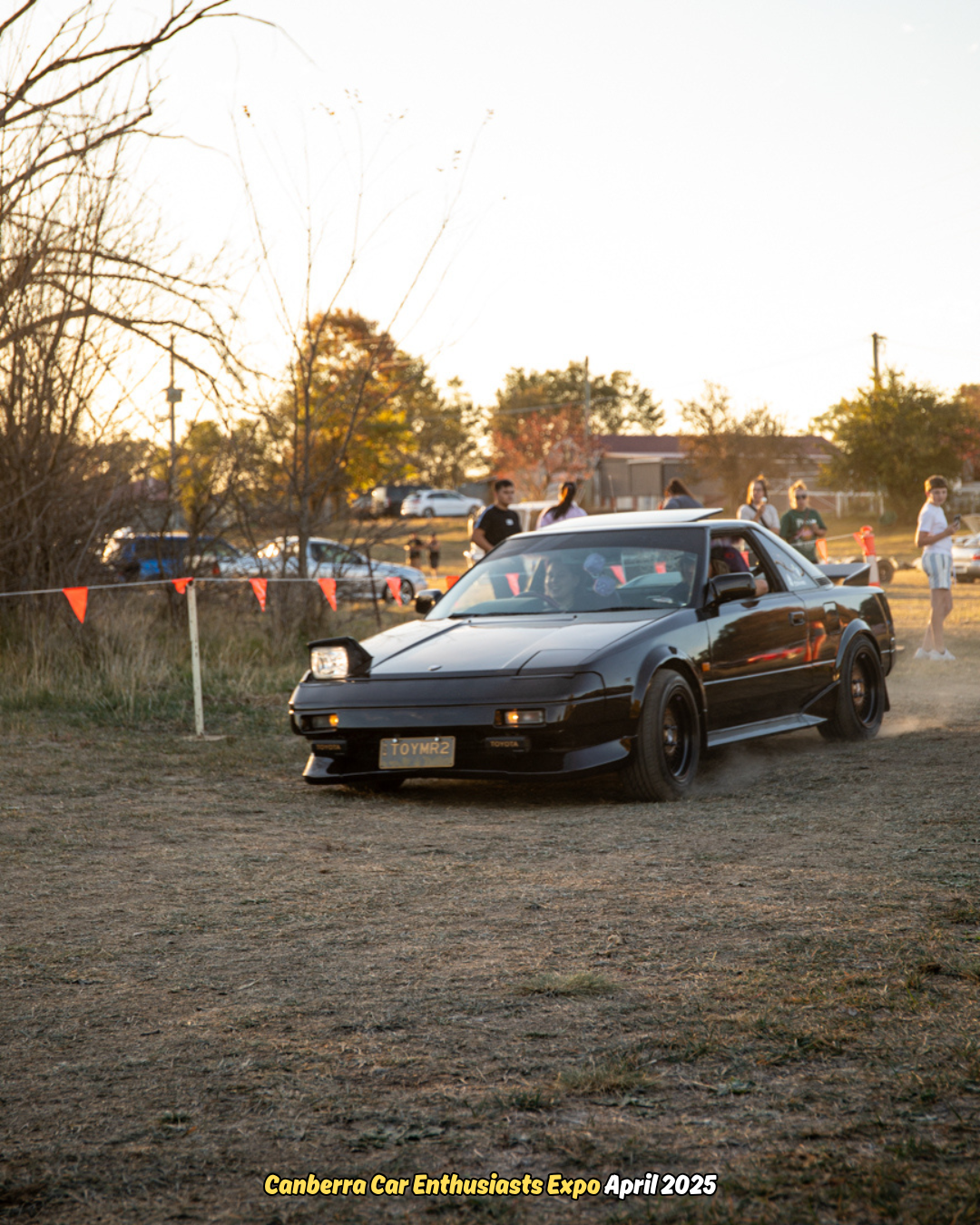 Black Toyota MR2 car driving on dirt at a car expo with people in the background.