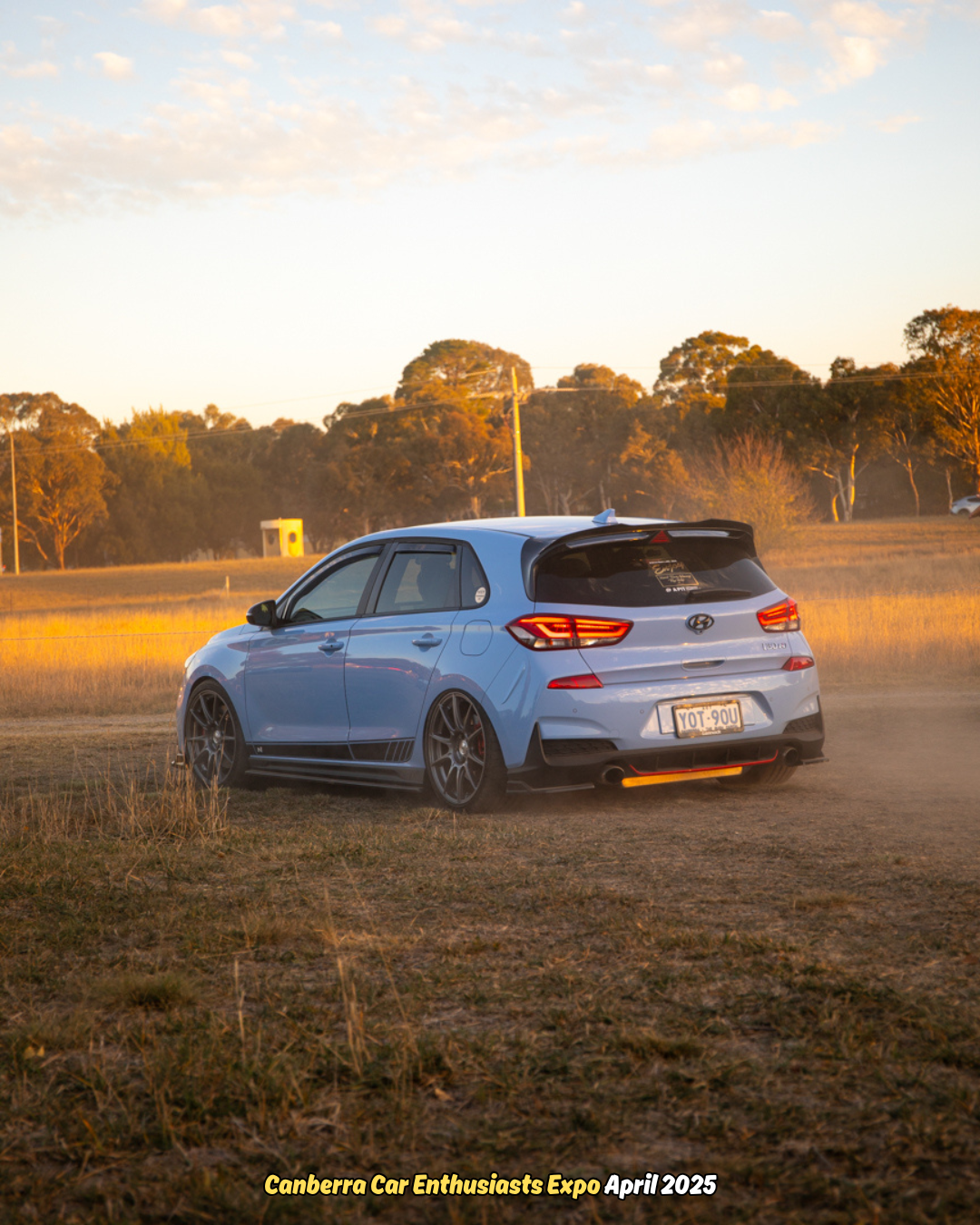Light blue Hyundai hatchback driving on a dirt road in a rural field at sunset.