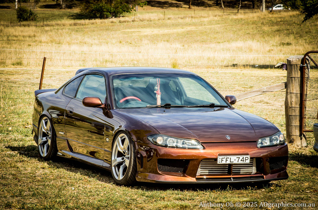 Brown modified sports car parked on grass near a wire fence in a rural area.