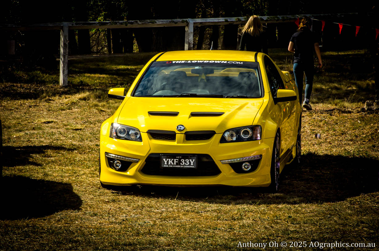 Bright yellow sports car parked on grass with two people walking in the background.