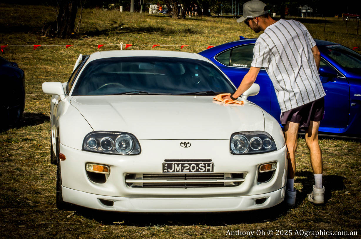 Man wearing a cap and striped shirt cleaning a white Toyota Supra at an outdoor car event.