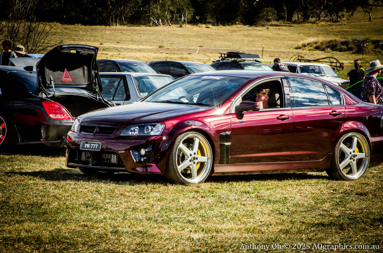 Shiny maroon sports sedan with large silver rims parked on grass at a car meet.