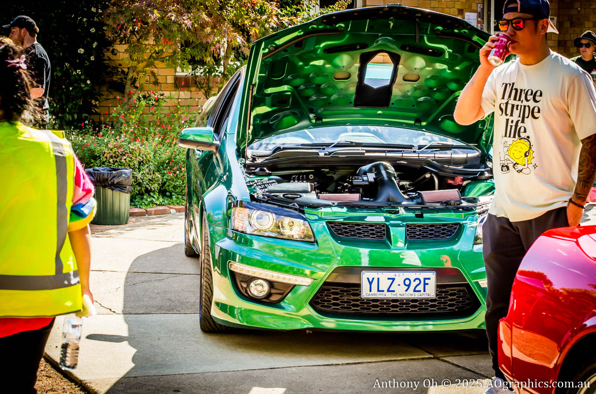 Green car with open hood on display at outdoor event with people nearby.