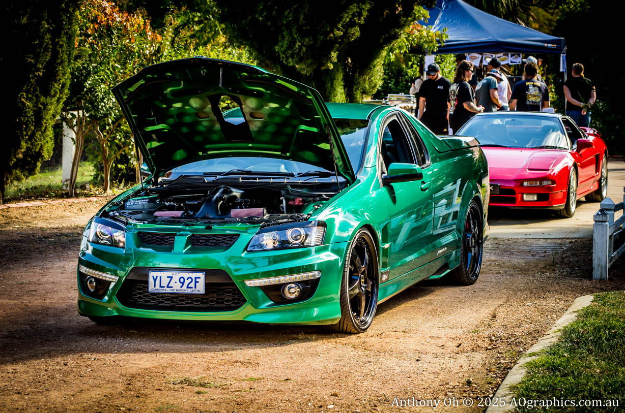 Green car with hood open and red sports car behind, people near food stall in background.
