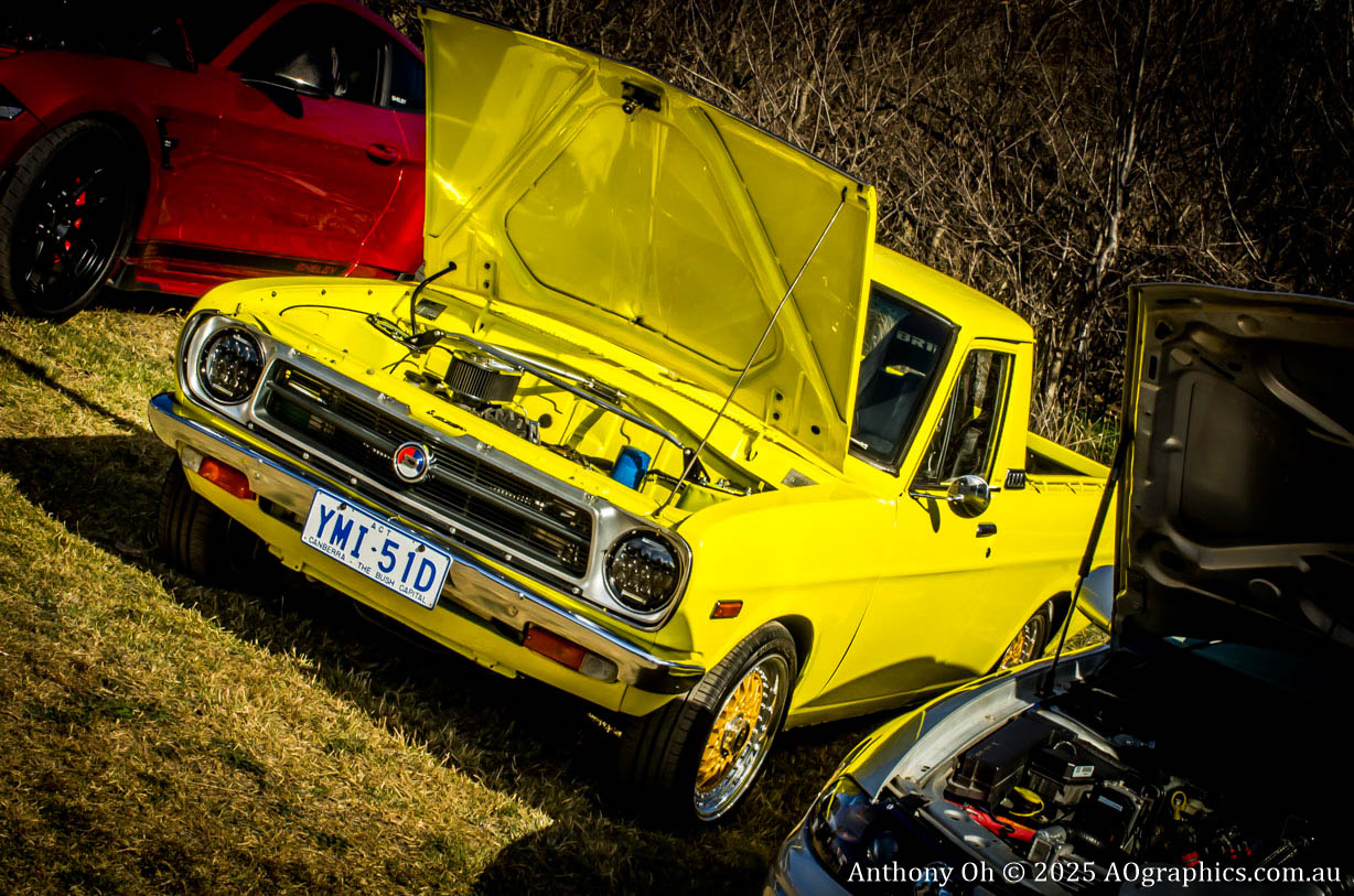 Yellow vintage pickup truck with open hood at a car show, grass and trees in background.