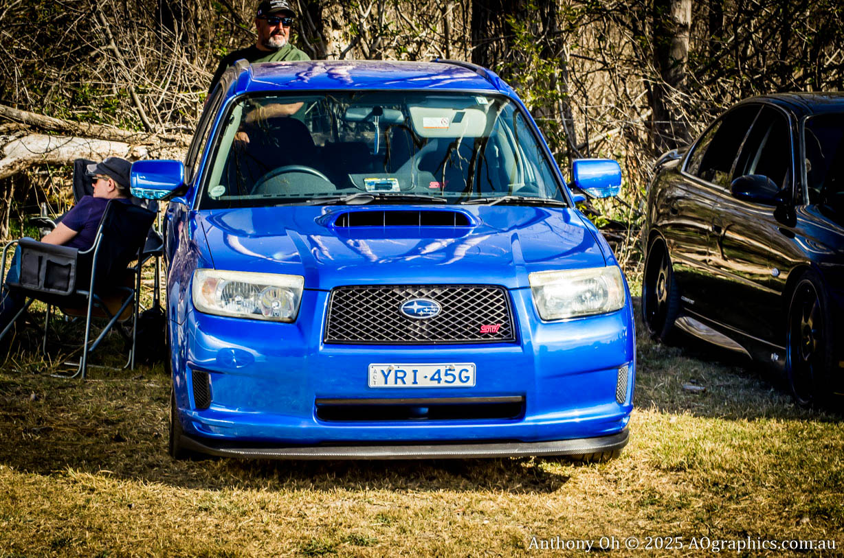 Blue Subaru WRX STI parked on grass with two men sitting nearby in a wooded area.