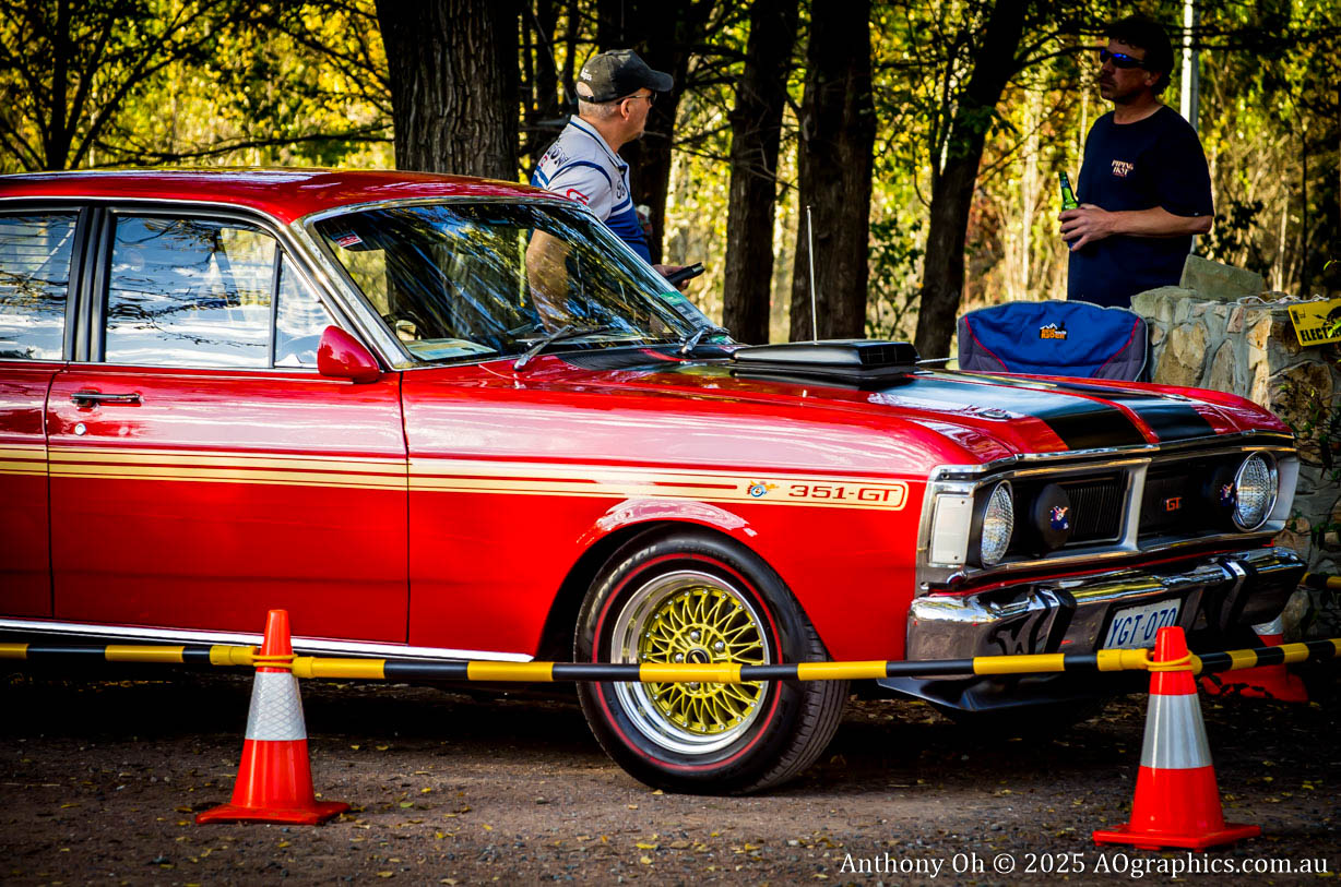 Red classic Ford 351 GT car parked near two men and traffic cones in a wooded area.