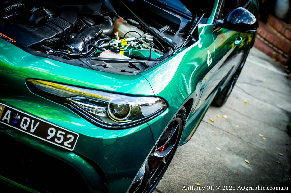 Close-up of a green sports car with hood open showing engine, parked on a concrete driveway.