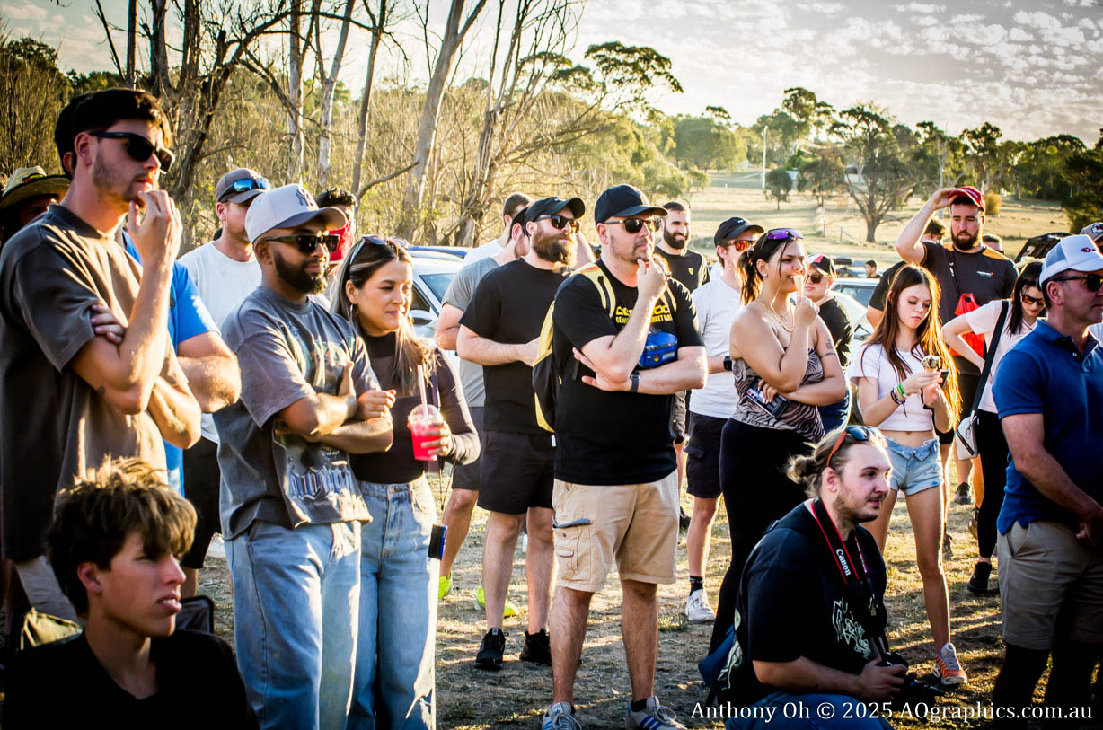 Group of people outdoors listening attentively, surrounded by trees and parked cars.