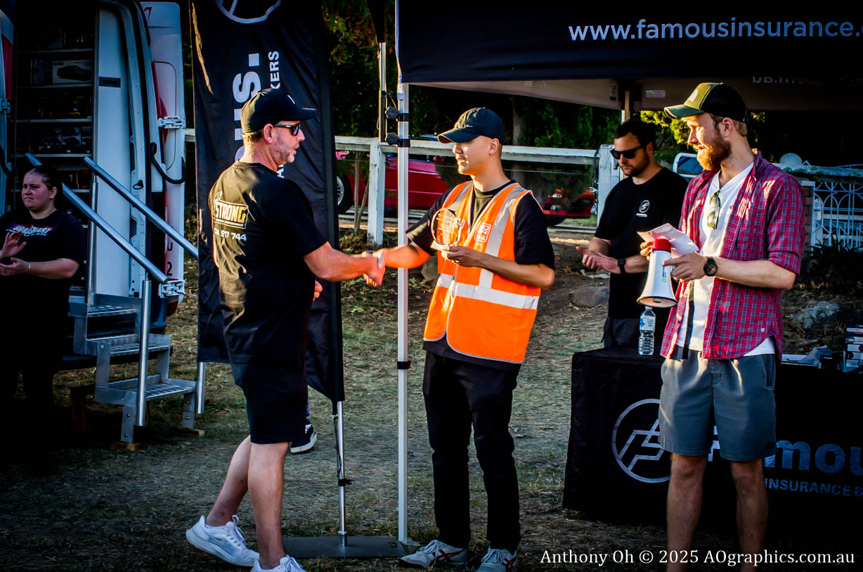 Two men shaking hands during an outdoor event, one wearing an orange safety vest and holding an award.