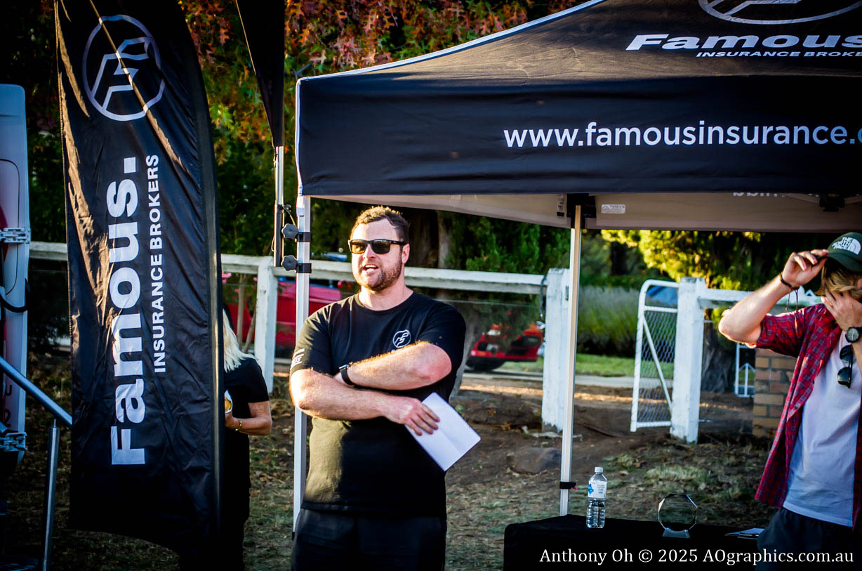 Man in sunglasses stands with arms crossed under Famous Insurance tent at outdoor event.