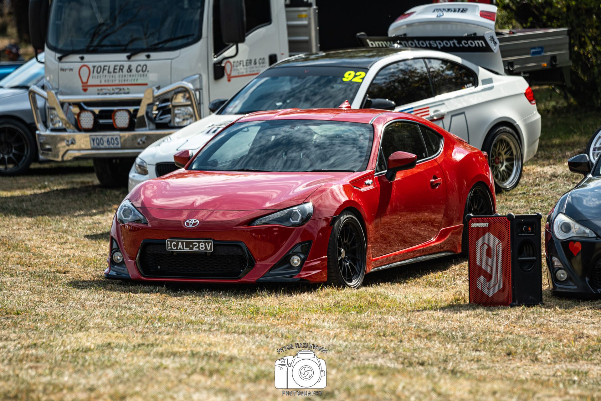 Red sports car parked on grass with a white rally car and other vehicles in the background.