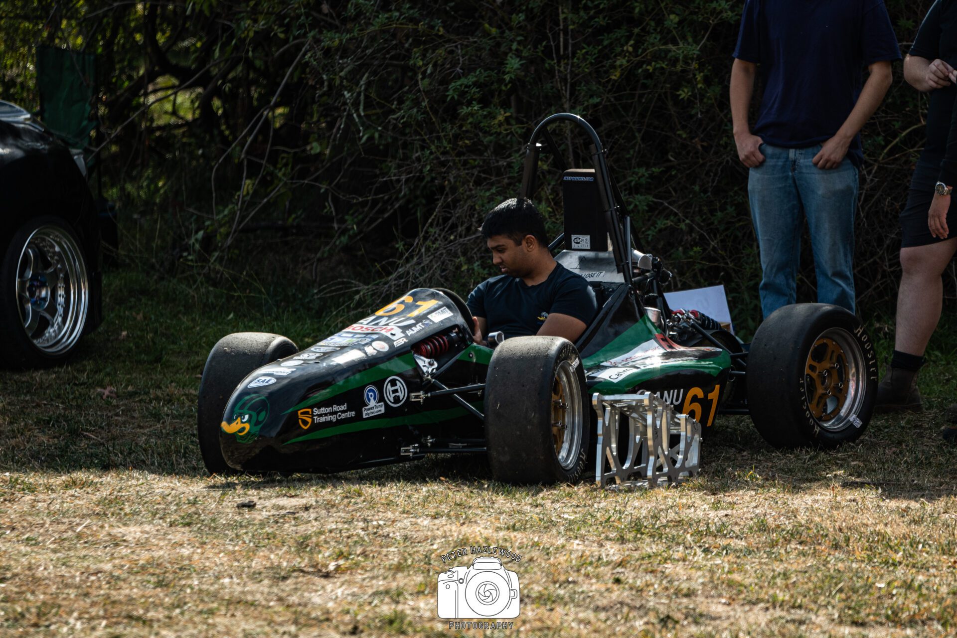 Man sitting in a small black race car with number 61 on grass beside two standing people.