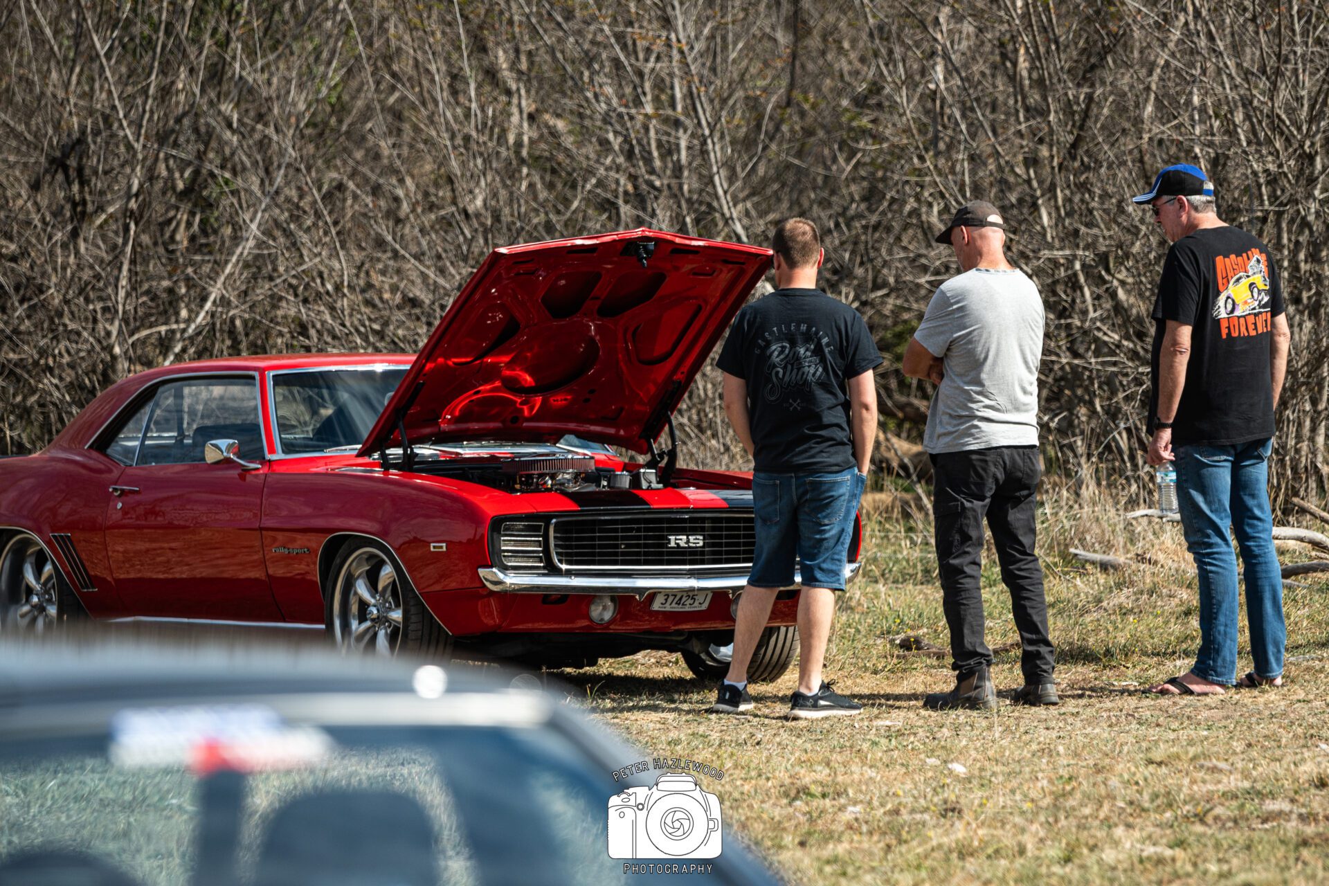 Three men examining a red vintage car with its hood open in an outdoor setting.