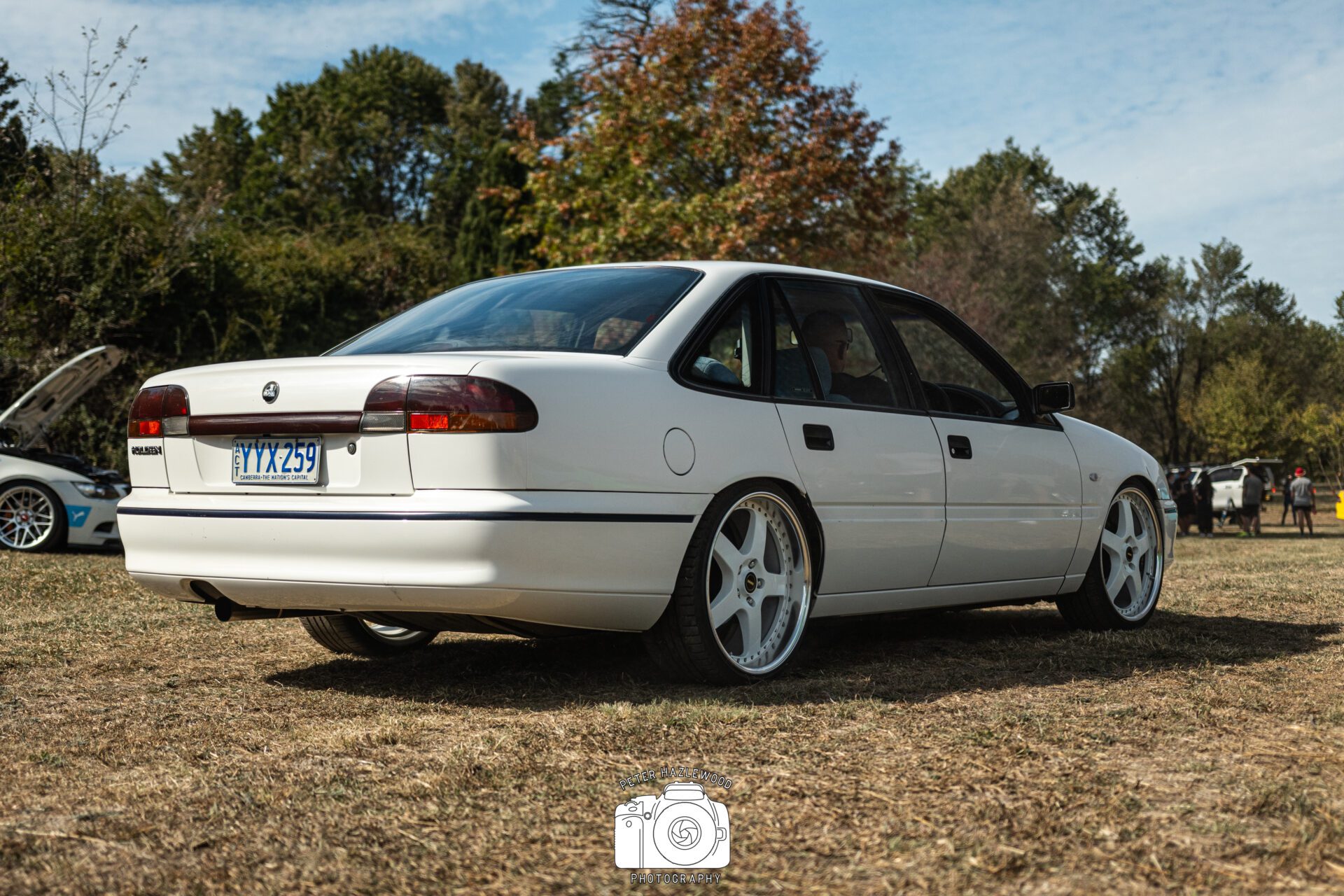 White Holden sedan with large aftermarket wheels parked on dry grass under a partly cloudy sky.