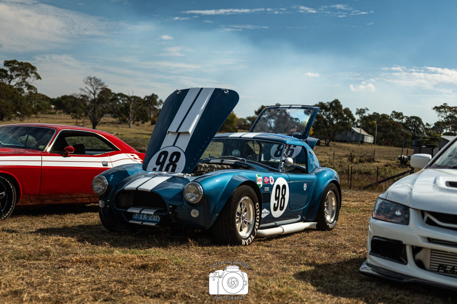 Blue vintage race car with number 98, hood and trunk open, parked on grass with other cars.
