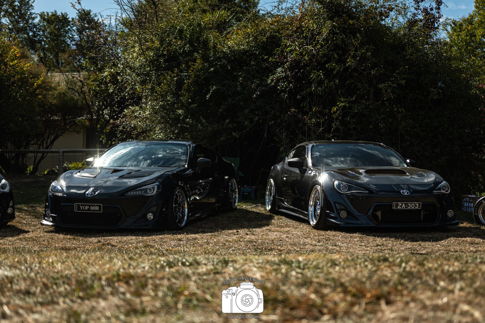 Two sleek black Toyota sports cars parked on grass with dense greenery in the background.