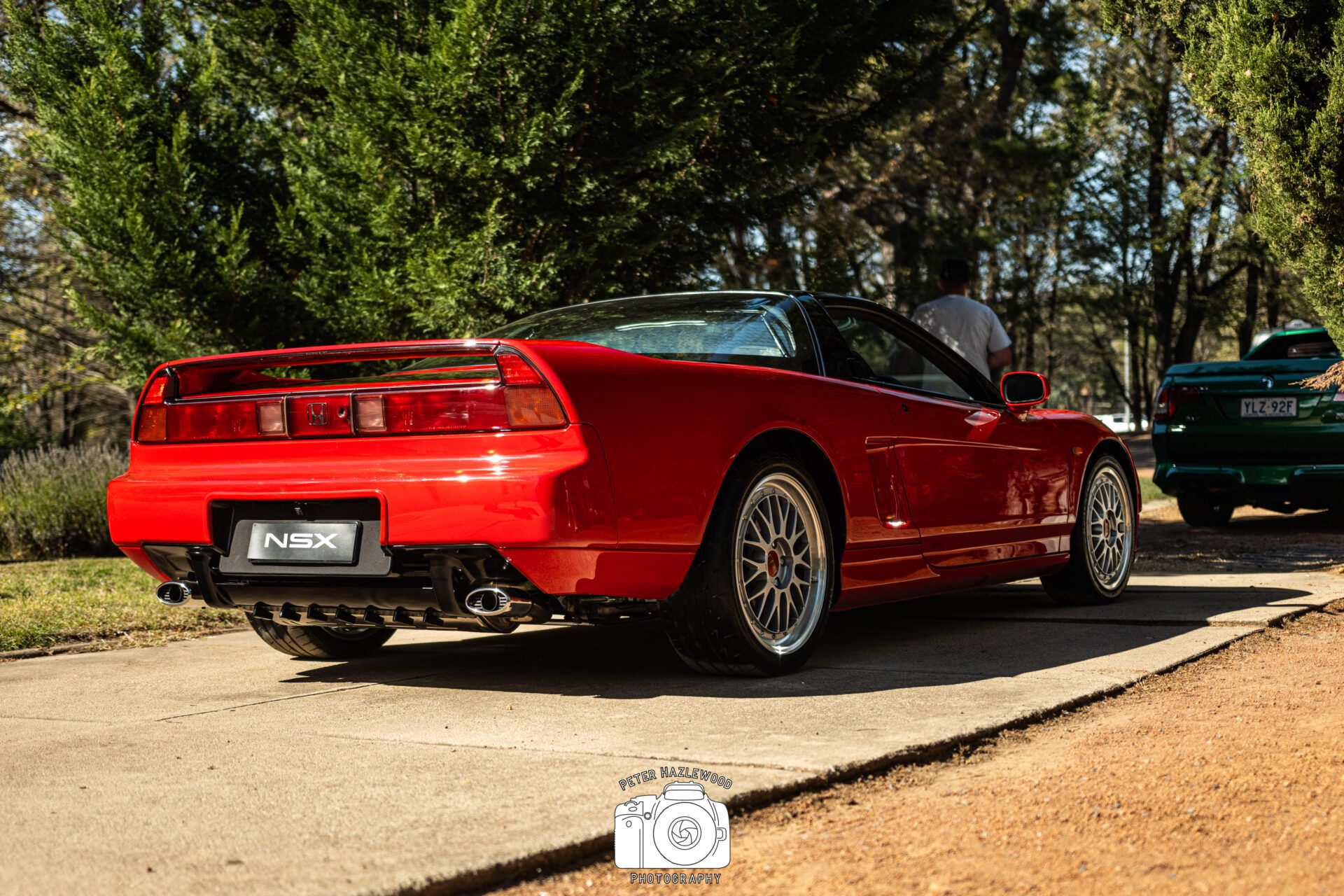 Red Honda NSX sports car parked on a driveway with trees and another car in the background.