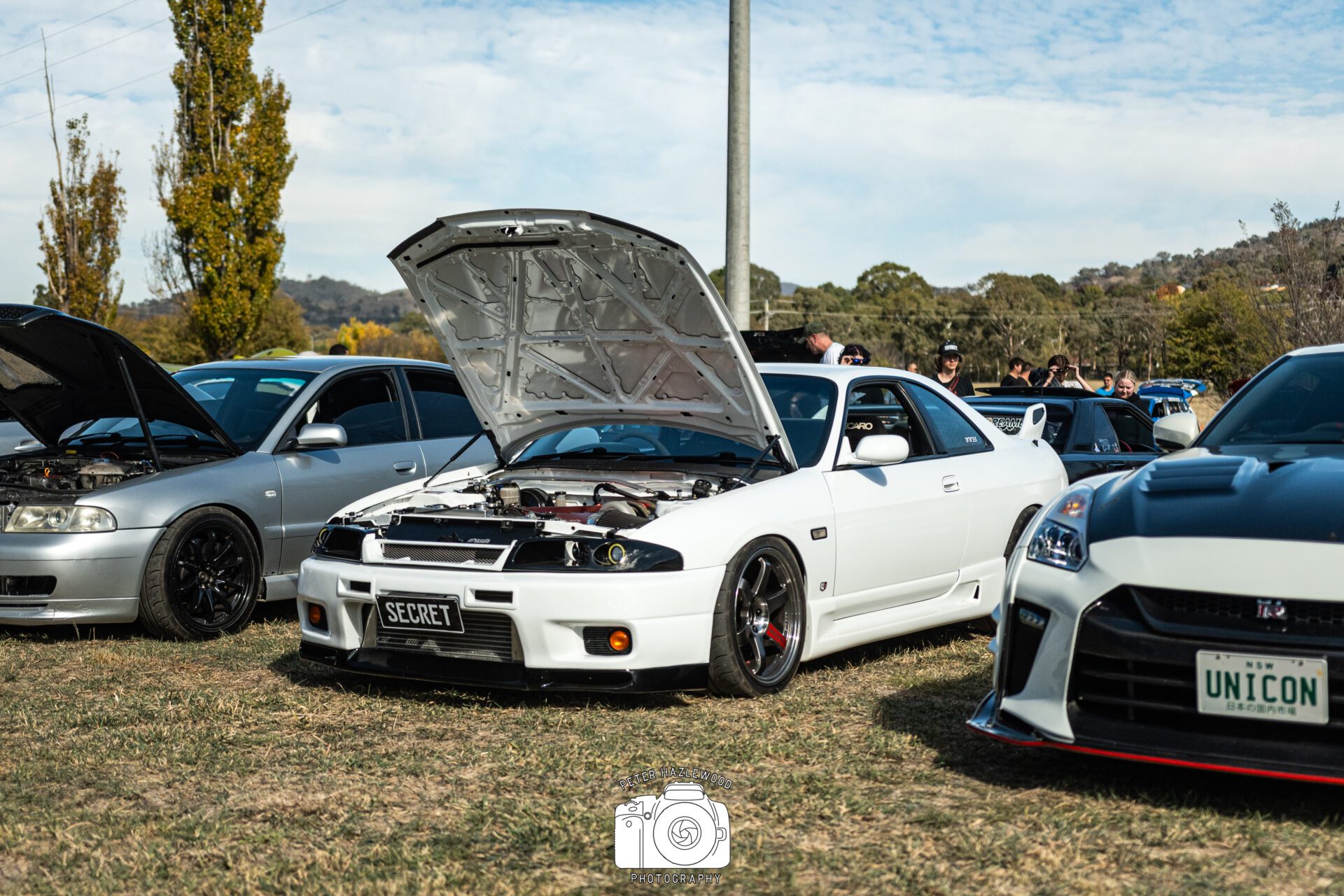 White sports car with open hood and "SECRET" plate, parked next to other modified cars at outdoor event.
