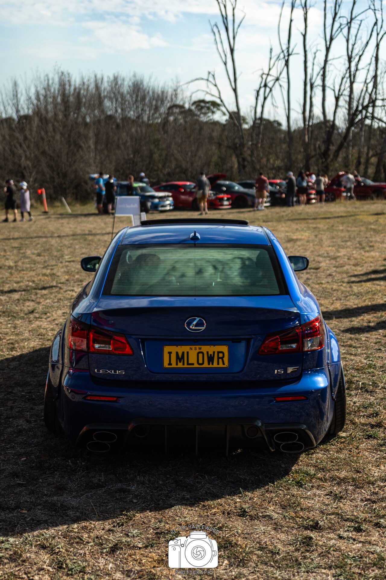 Rear view of a blue Lexus IS F with custom license plate "IMLOWR" at outdoor car meet.