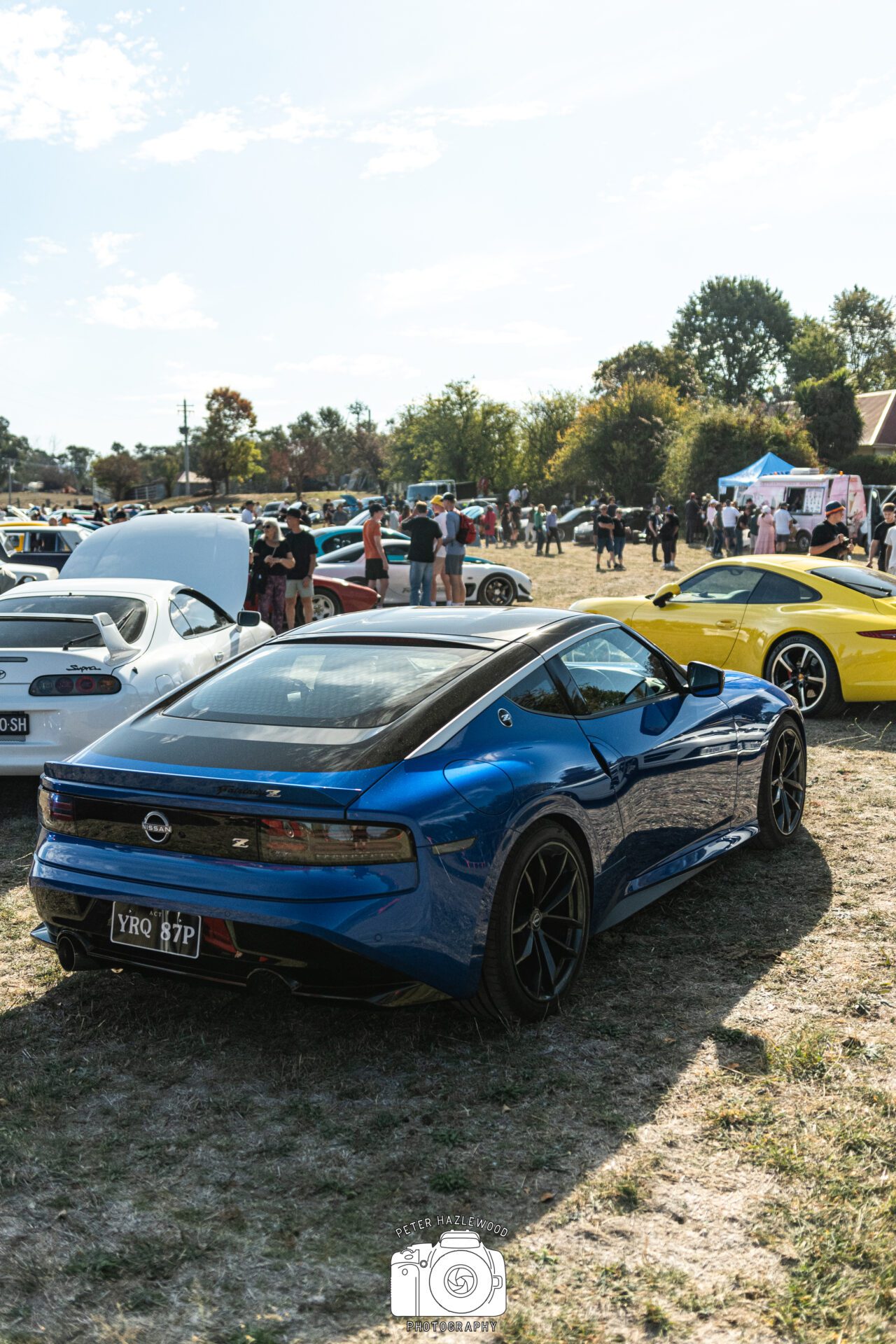 Blue Nissan Fairlady Z parked on grass at car meet with other sports cars and people in background.